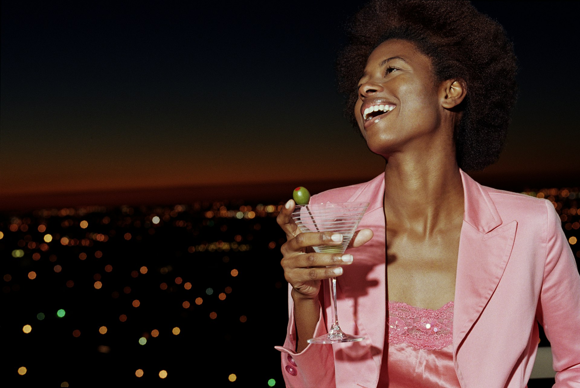 A woman wearing a pink jacket over a shiny pink top holds a martini glass while smiling on a balcony at night.