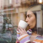 A woman looks out of a window while sipping from a white coffee mug. 