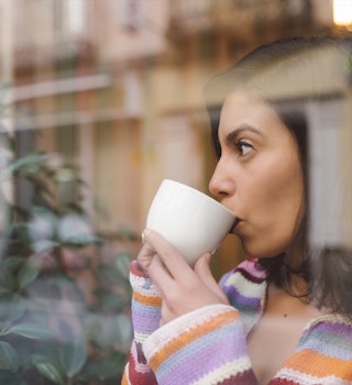 A woman looks out of a window while sipping from a white coffee mug. 
