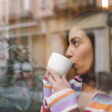 A woman looks out of a window while sipping from a white coffee mug.