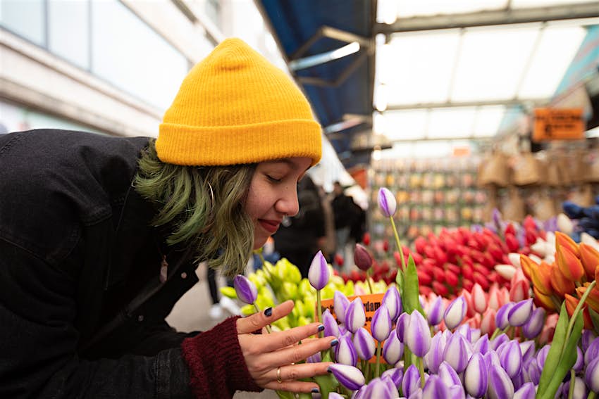 A woman wearing a yellow hat bends down to smell a bouquet of tulips