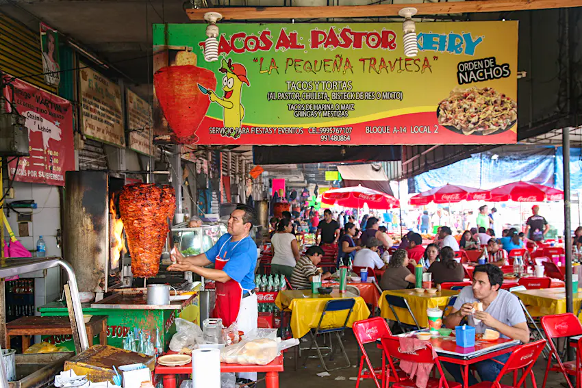 Restaurants serving quick meals at Mercado Municipal Lucas de Gálvez in Mérida, Mexico A man carves meat for tacos al pastor among crowded tables at the Mercado Municipal Lucas de Gálvez market in Mérida