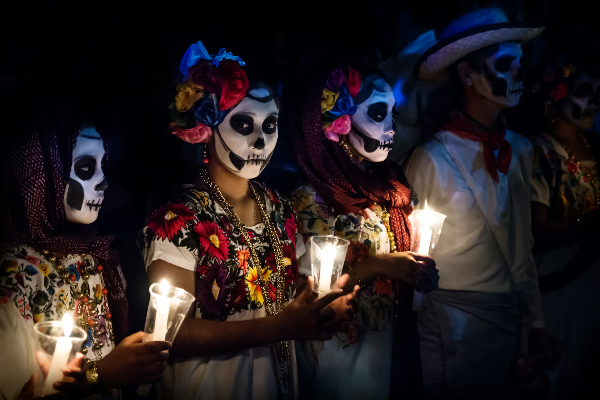 Three women with Catrina costumes and a man in white cowboy dress, wearing skull makeup and holding candles at a Dia de Muertos parade