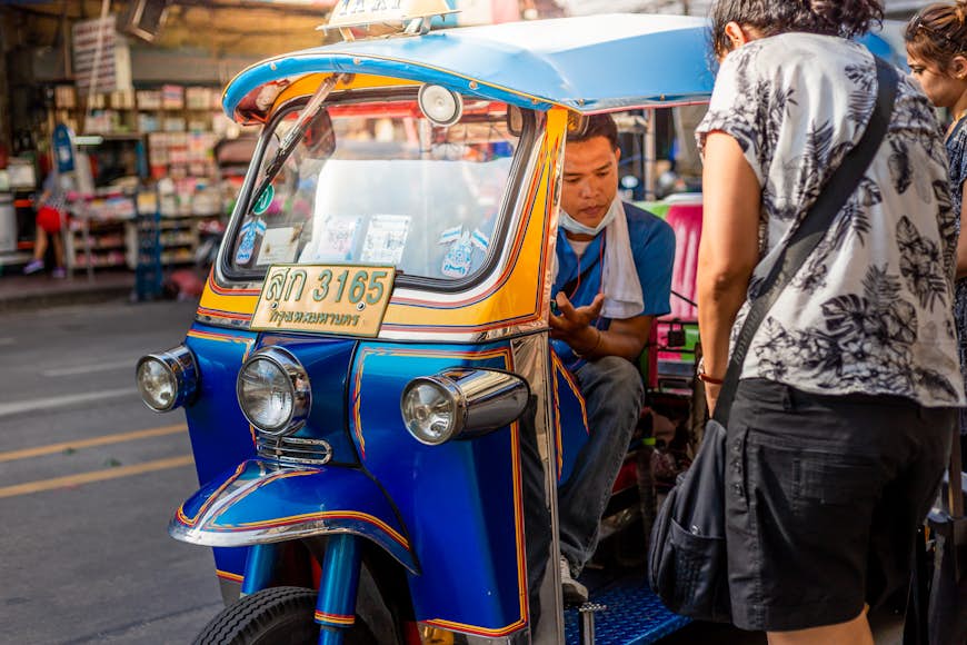A driver in a blue and white tuk-tuk, picking up a passenger