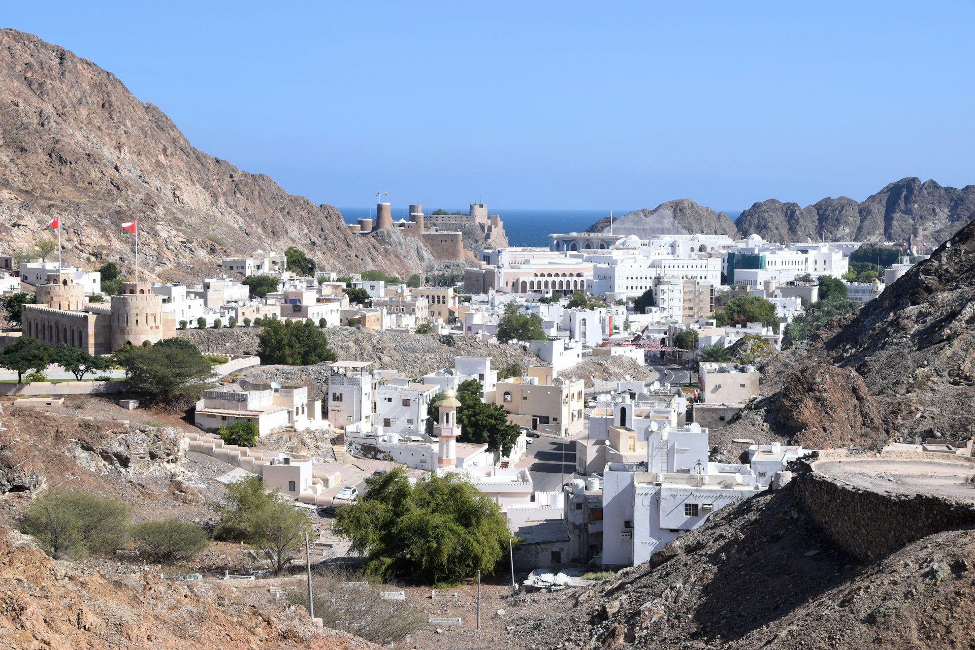View of the white buildings of the old town of Muscat, Oman, among brown mountains and the blue Gulf of Oman in the distance