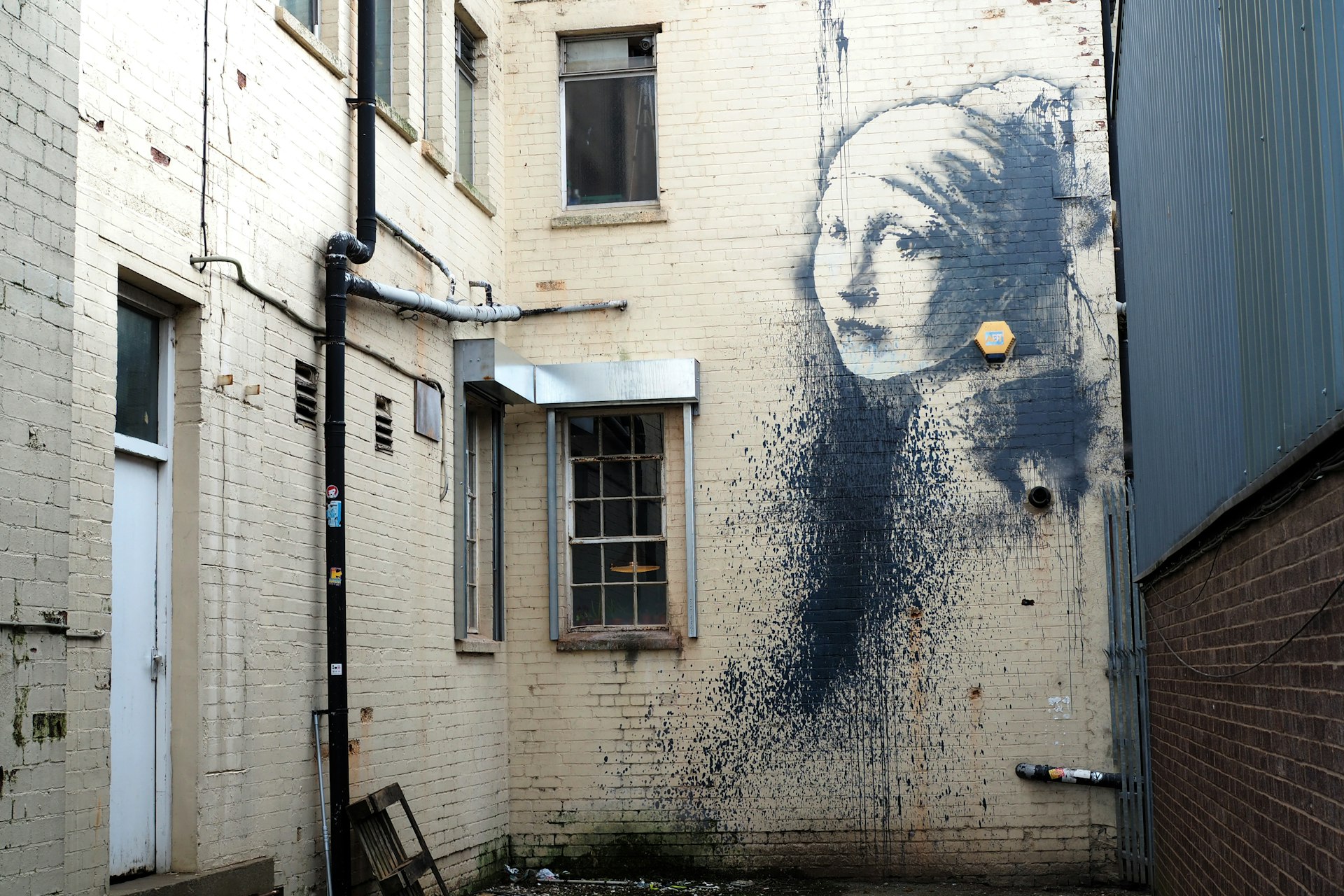 A white brick wall in an alley showing “Girl with Pierced Eardrum” by street artist Banksy