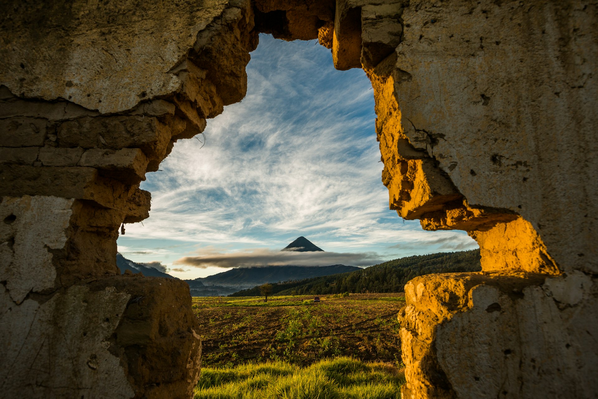 A cone-shaped volcano topped with clouds, framed by a broken stone wall in Quetzaltenango, Guatemala. 