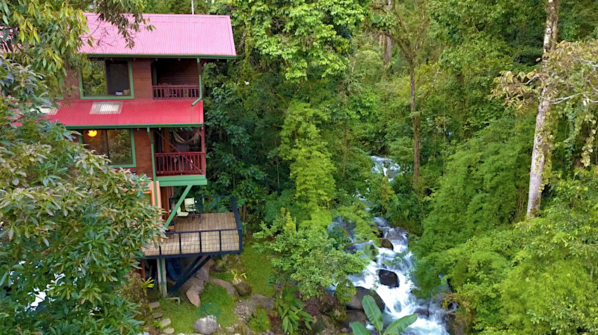 Vista exterior de un AirBNB de varios niveles con techo rojo en medio de una selva tropical y junto a un arroyo de rápido movimiento en Costa Rica. 