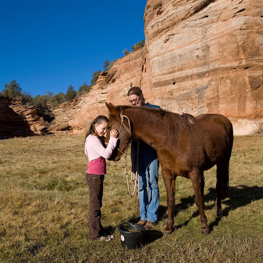 A girl feeds a horse in front of a rock formation and a bright blue sky at Best Friends Animal Sanctuary, Utah