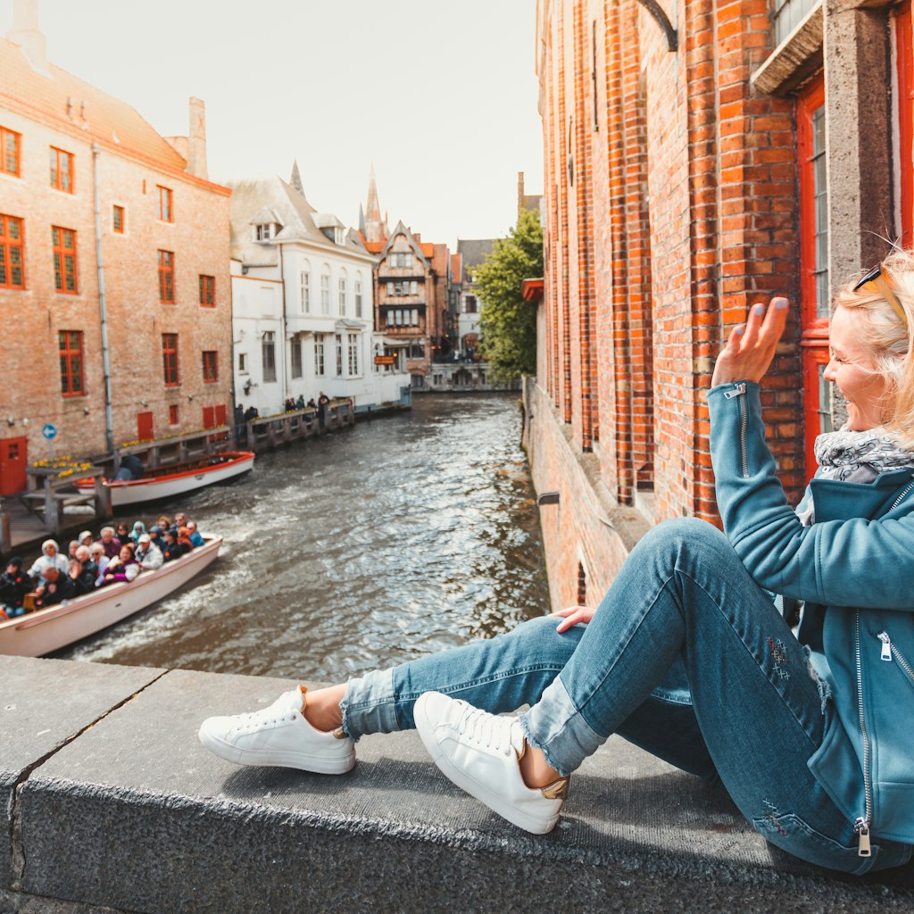Young woman tourist sits on a bridge in the old city in Bruges in the morning in Belgium and waves a hand passing by a tourist boat with tourists.; Shutterstock ID 1400041118; your: Claire Naylor; gl: 65050; netsuite: Online editorial; full: Bruges things to do