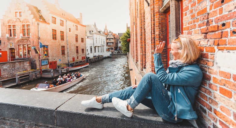 Young woman tourist sits on a bridge in the old city in Bruges in the morning in Belgium and waves a hand passing by a tourist boat with tourists.; Shutterstock ID 1400041118; your: Claire Naylor; gl: 65050; netsuite: Online editorial; full: Bruges things to do
