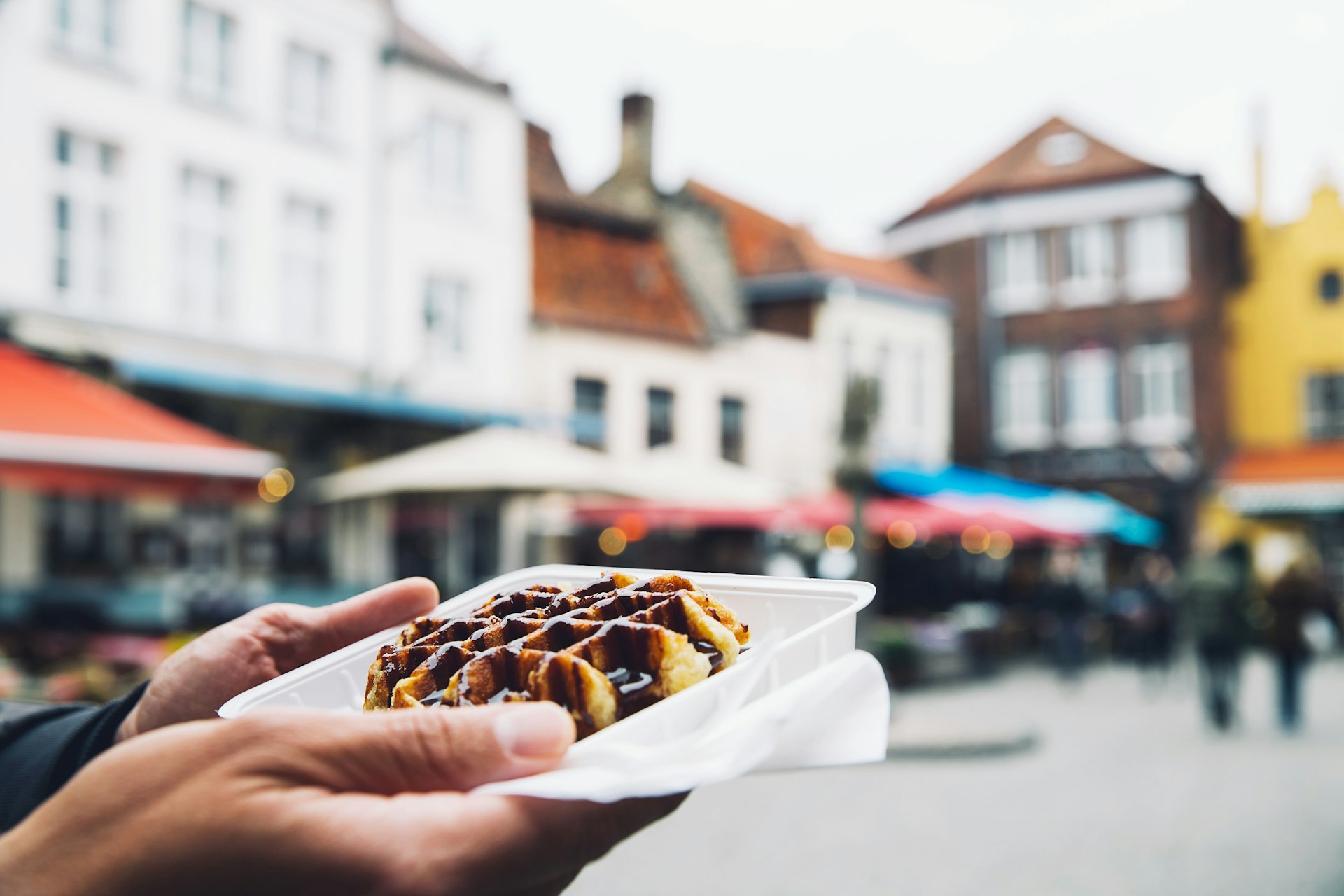A person holds a takeaway tray containing a waffle covered in chocolate sauce outside in a medieval square. 