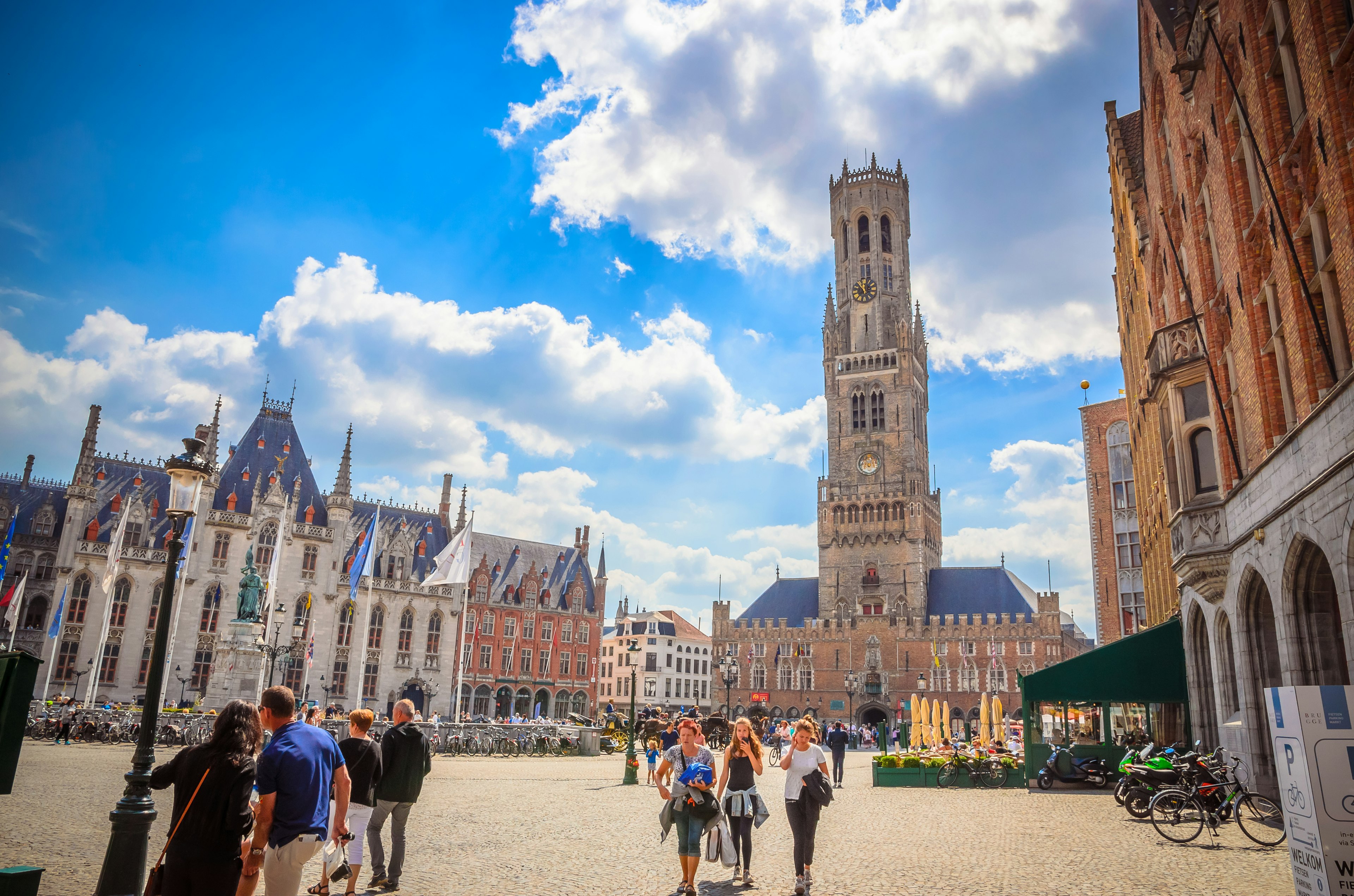 People walk through a huge medieval square dominated by a tall Gothic tower