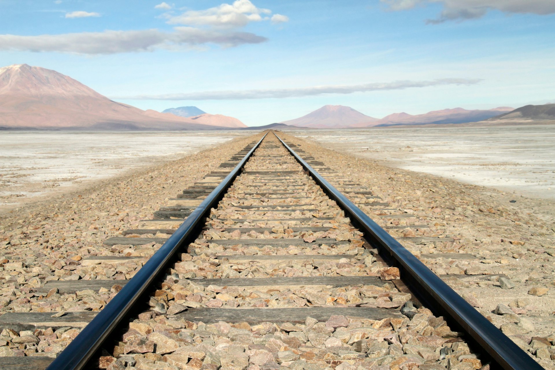 Rail lines stretch off straight into the distance across white salt flats surrounded by red-brown mountains