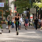 A man and a woman ride motorized scooters down the sidewalk of downtown Charlotte. There are a group of people also walking down the sidewalk. 