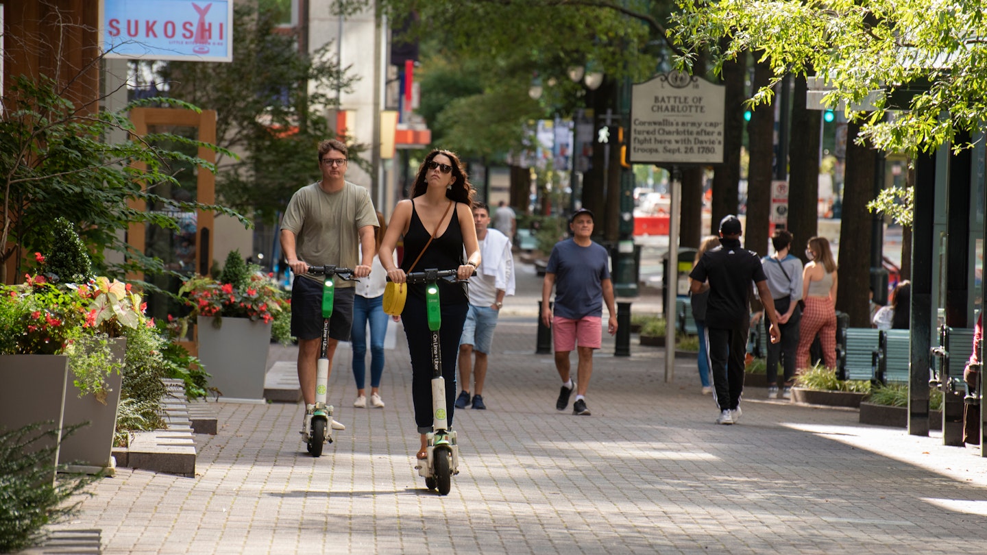 A man and a woman ride motorized scooters down the sidewalk of downtown Charlotte. There are a group of people also walking down the sidewalk. 
