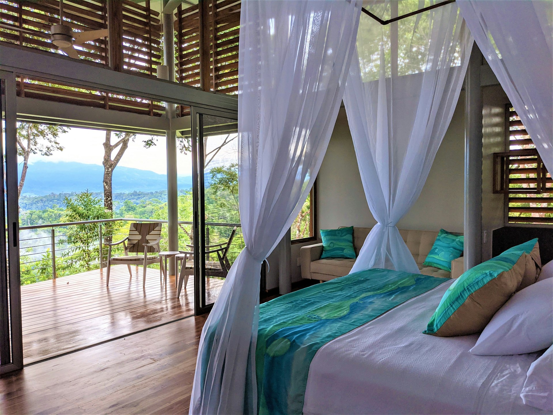 Interior shot of the inside of the Cielo Lodge. There's a bed with white curtains draping from the bed frame and a sofa with bright sea green pillows. The balcony offers views lush country. 