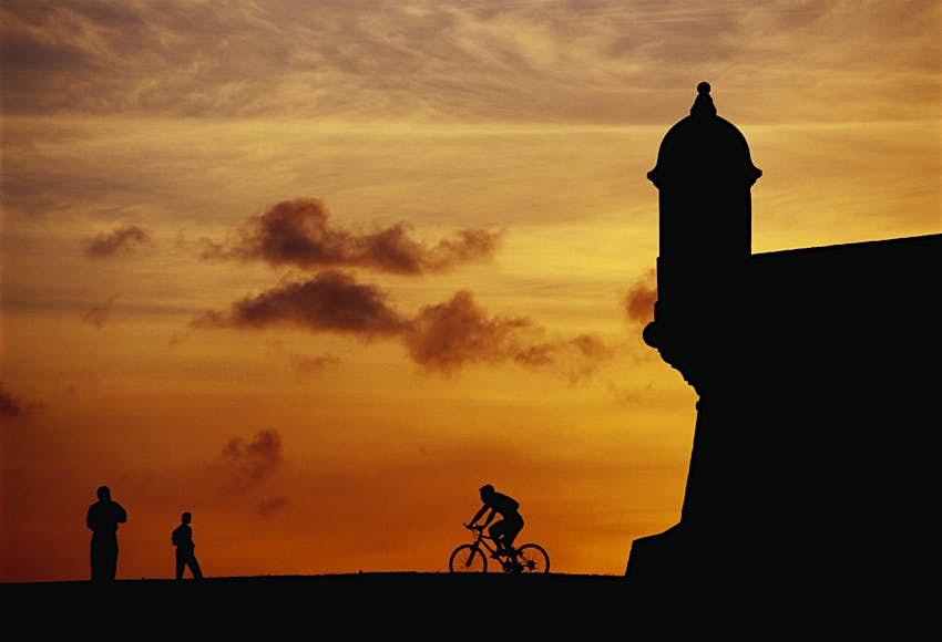A trio of people, including one on a bicycle, in silhouette alongside Fort El Morro in San Juan, Puerto Rico. 