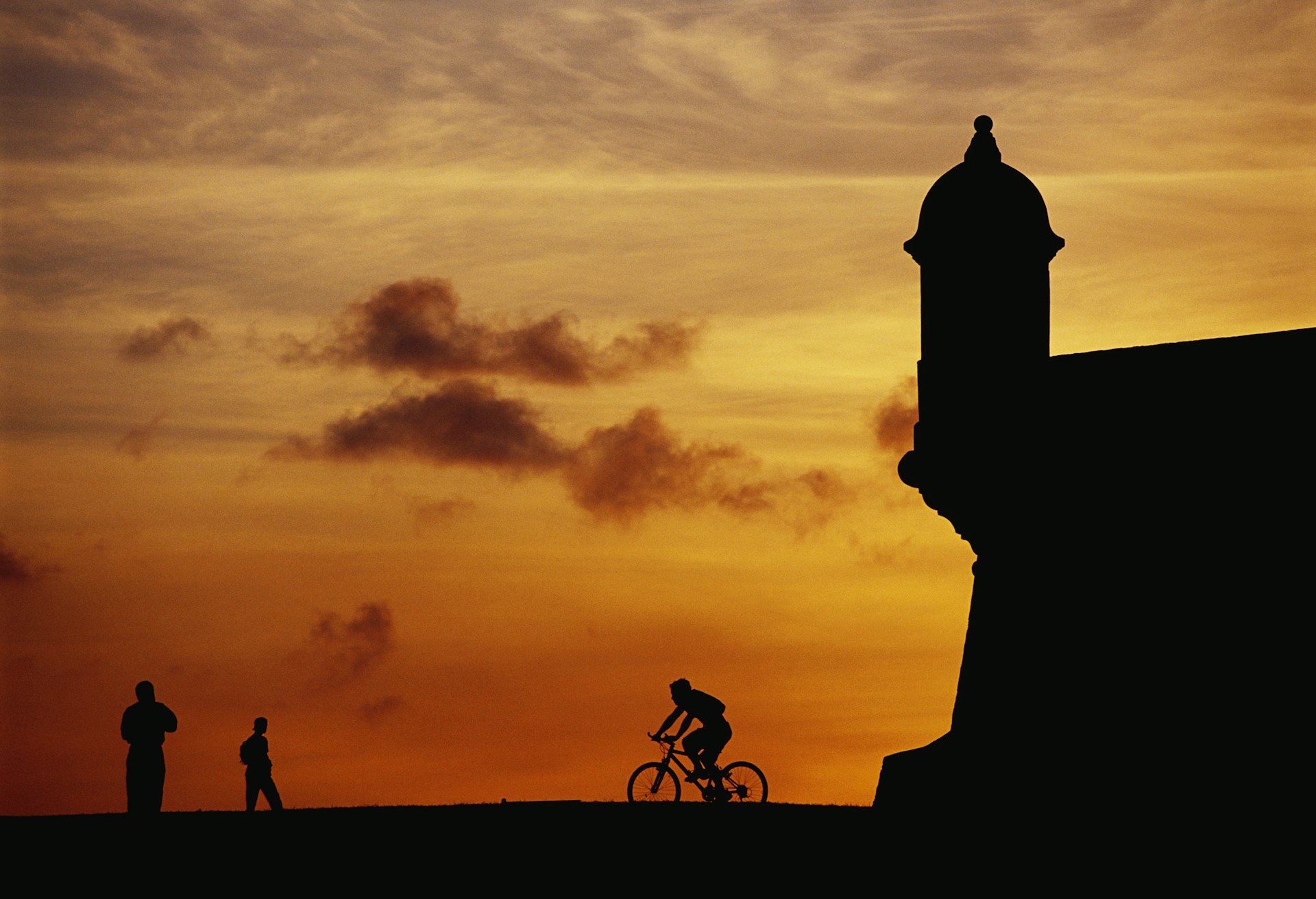 A trio of people, including one a bike, in silhouette alongside El Morro Fort in San Juan, Puerto Rico. 
