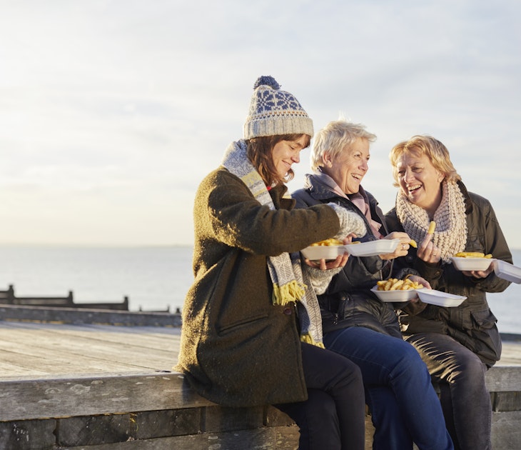 Three white women, one middle aged, two slightly older sit and smile on a sea groyne in 
Whitstable, Kent, England whilst eating takeaway fish and chips out of paper in warm, winter clothes.
