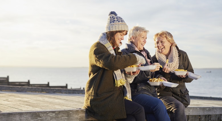 Three white women, one middle aged, two slightly older sit and smile on a sea groyne in 
Whitstable, Kent, England whilst eating takeaway fish and chips out of paper in warm, winter clothes.