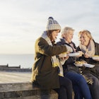 Three white women, one middle aged, two slightly older sit and smile on a sea groyne in
Whitstable, Kent, England whilst eating takeaway fish and chips out of paper in warm, winter clothes.
