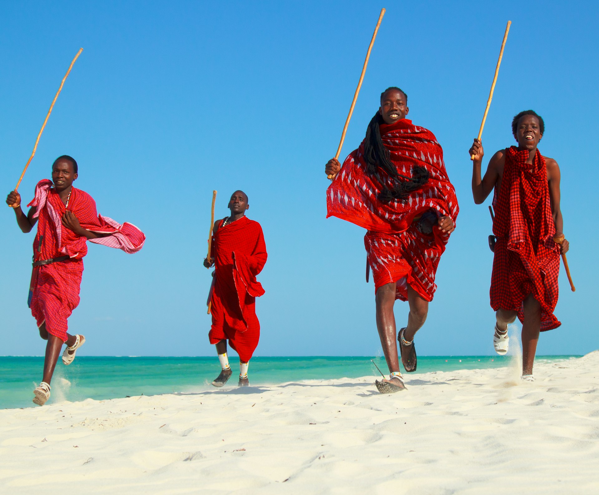 4 Maasai warriors, wearing red, holding sticks, jumping on a beach in Zanzibarr