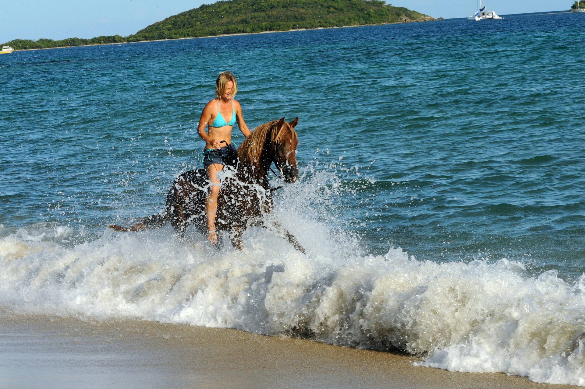 A woman smiles as she rides a horse through the crashing the surf on a beach