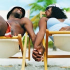 Two people hold hands and gaze at each other while sitting in comfortable beach chairs under palm trees. Camera angle is close on their entwined hands.