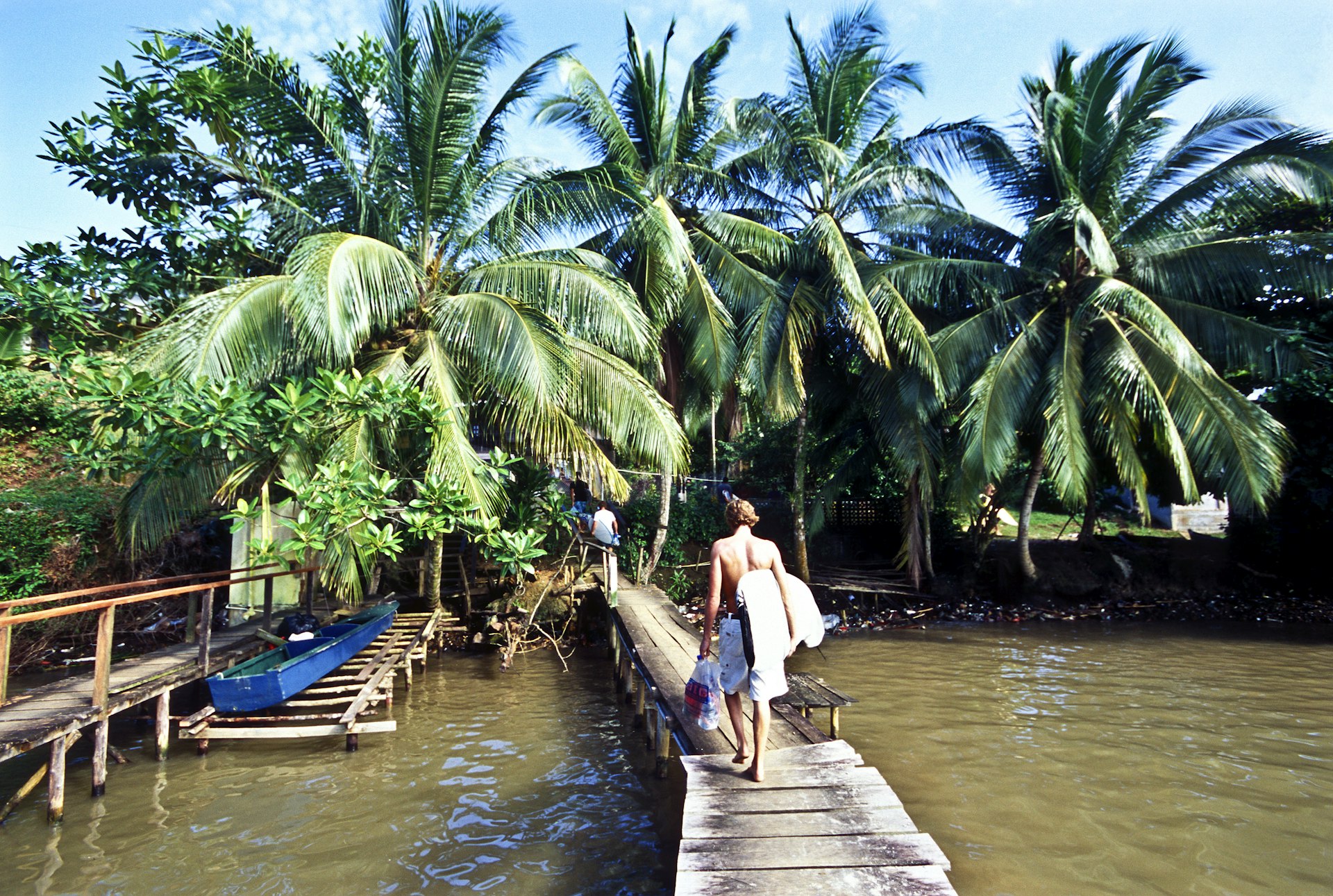 A man holding a surfboard under his arm walks down a dock toward a shore filled with palm trees in Bocas del Toro, Panama