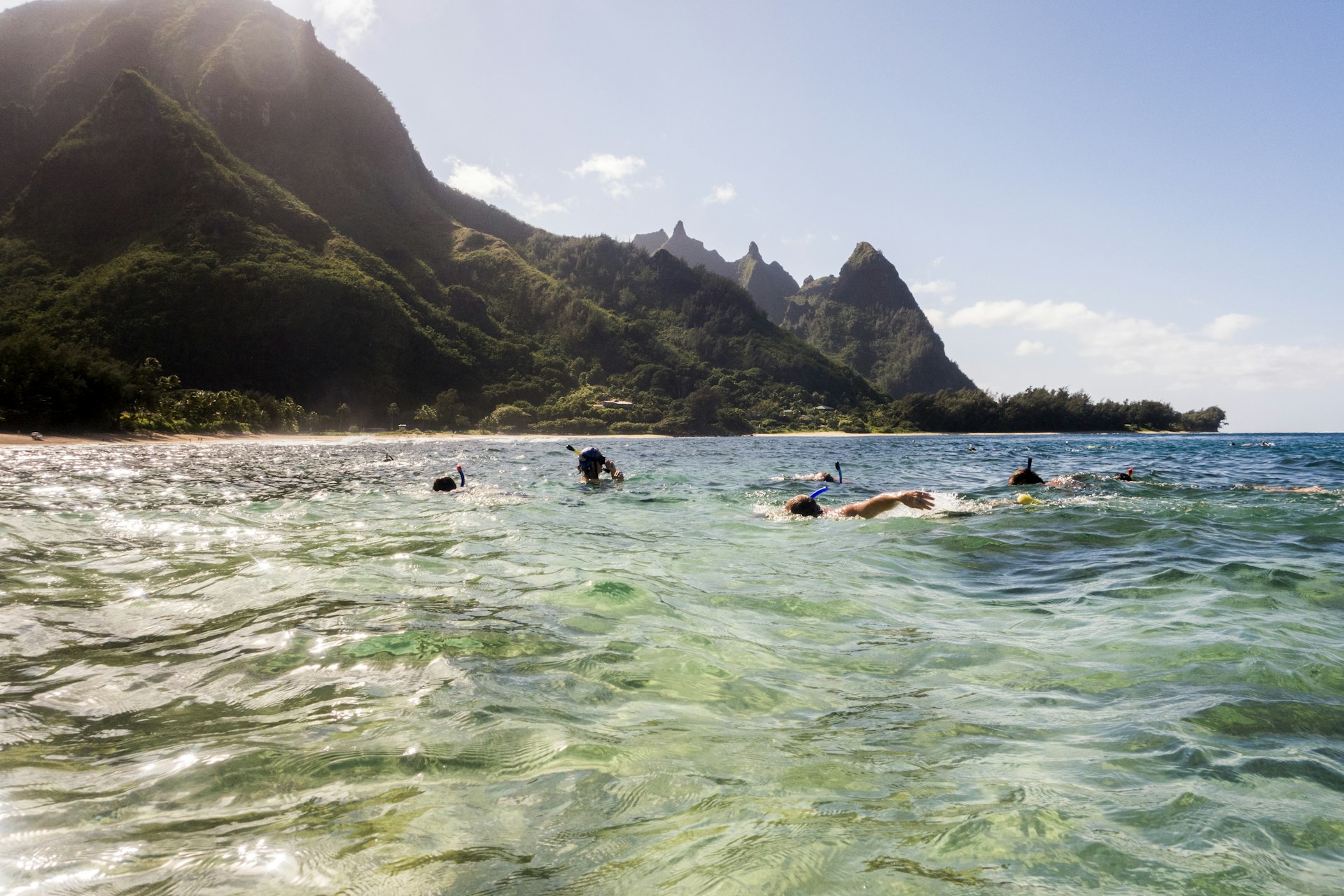 A group of people scuba diving and snorkeling off the coast of Kaua‘i on a sunny day with dramatic cliffs in the distance