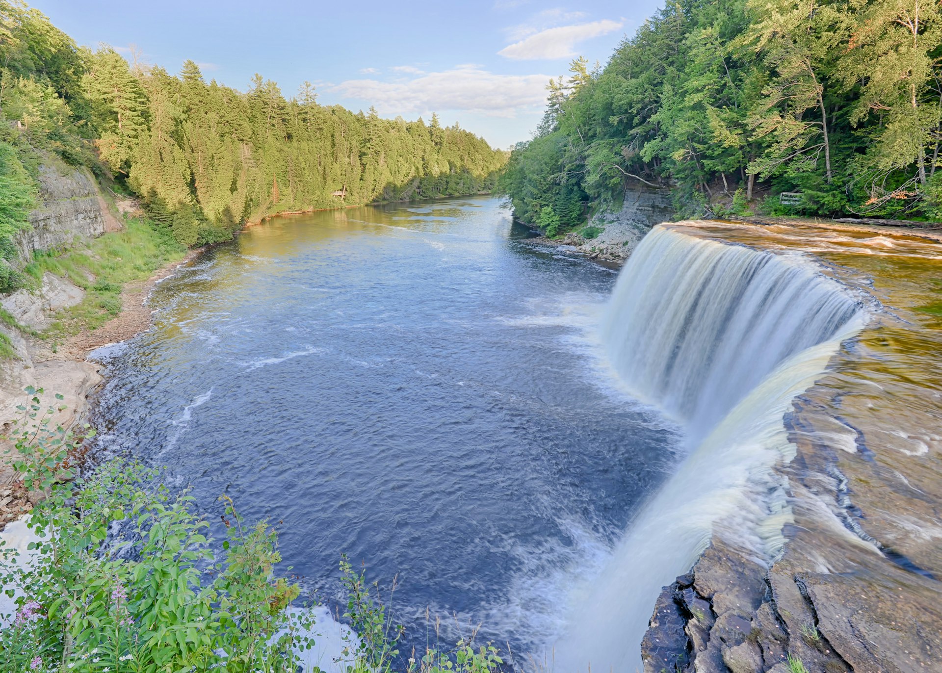 A curved waterfall flows into a river surrounded by woodland