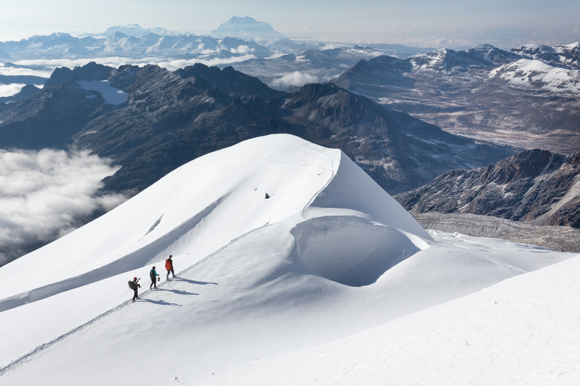 Three mountaineers on a snow ridge with cloud-covered mountains below, near or on Huayna Potosí