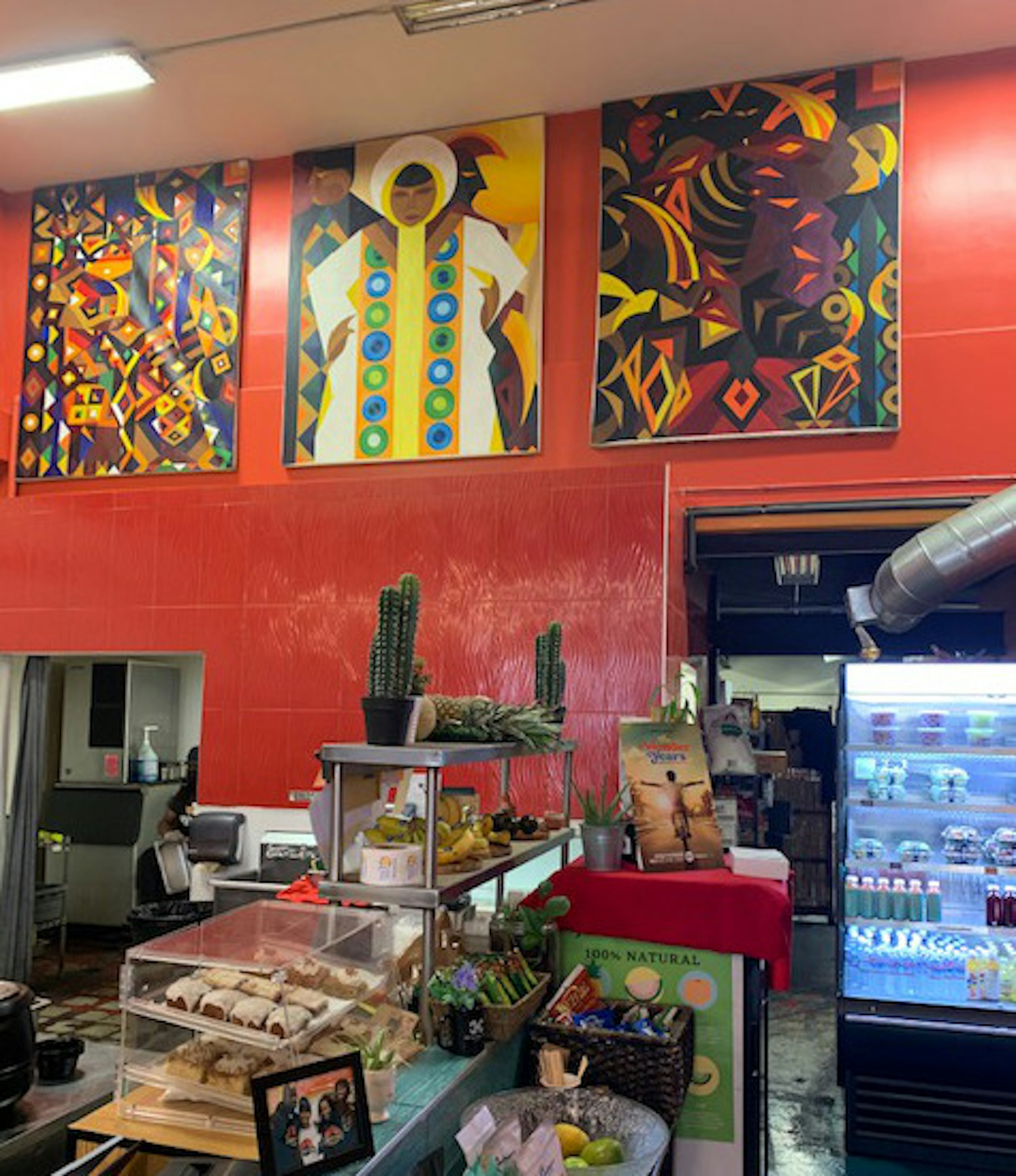 Interior shot of the Hot and Cool Cafe in Leimert Park. A trio of colorful paintings hang on the red wall. The counters are filled with sweet treats and fruits and off to the left is a refrigerator.