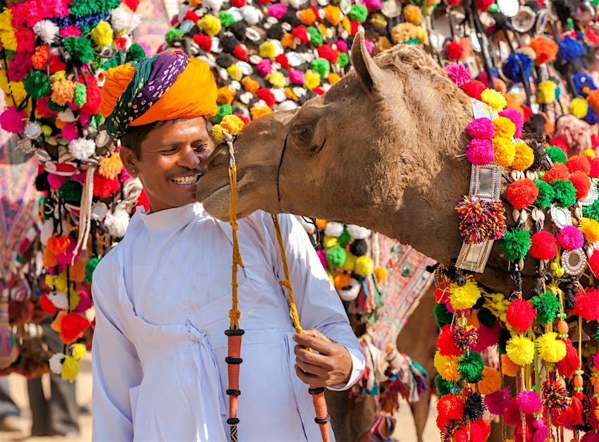 Ένας άντρας με τουρμπάνι χαμογελά καθώς μια καμήλα με βαριά διακόσμηση σπρώχνει τη μύτη του προς το πρόσωπό του