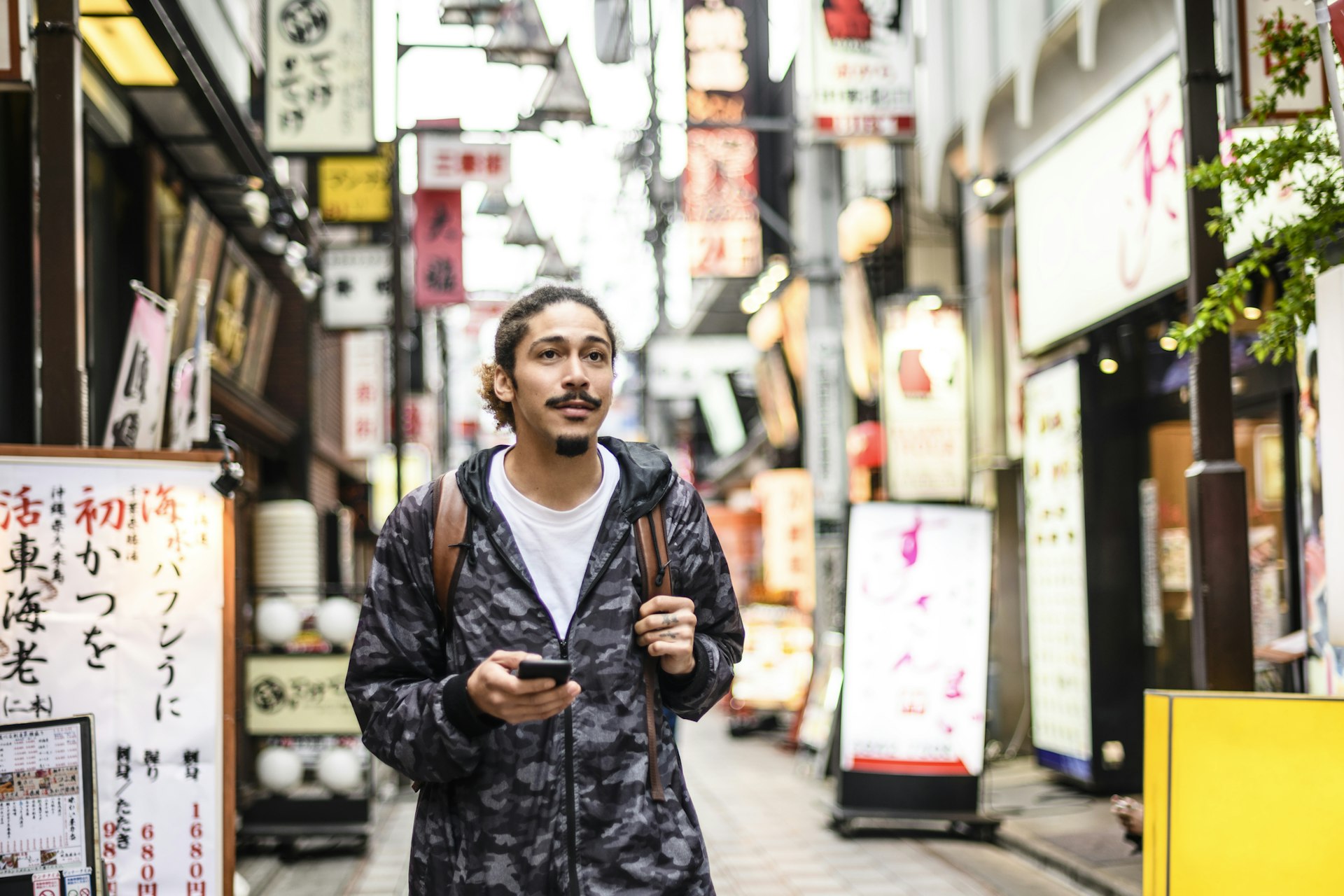 A young man walks down an urban street holding his phone. There are many Japanese signs on the buildings behind him