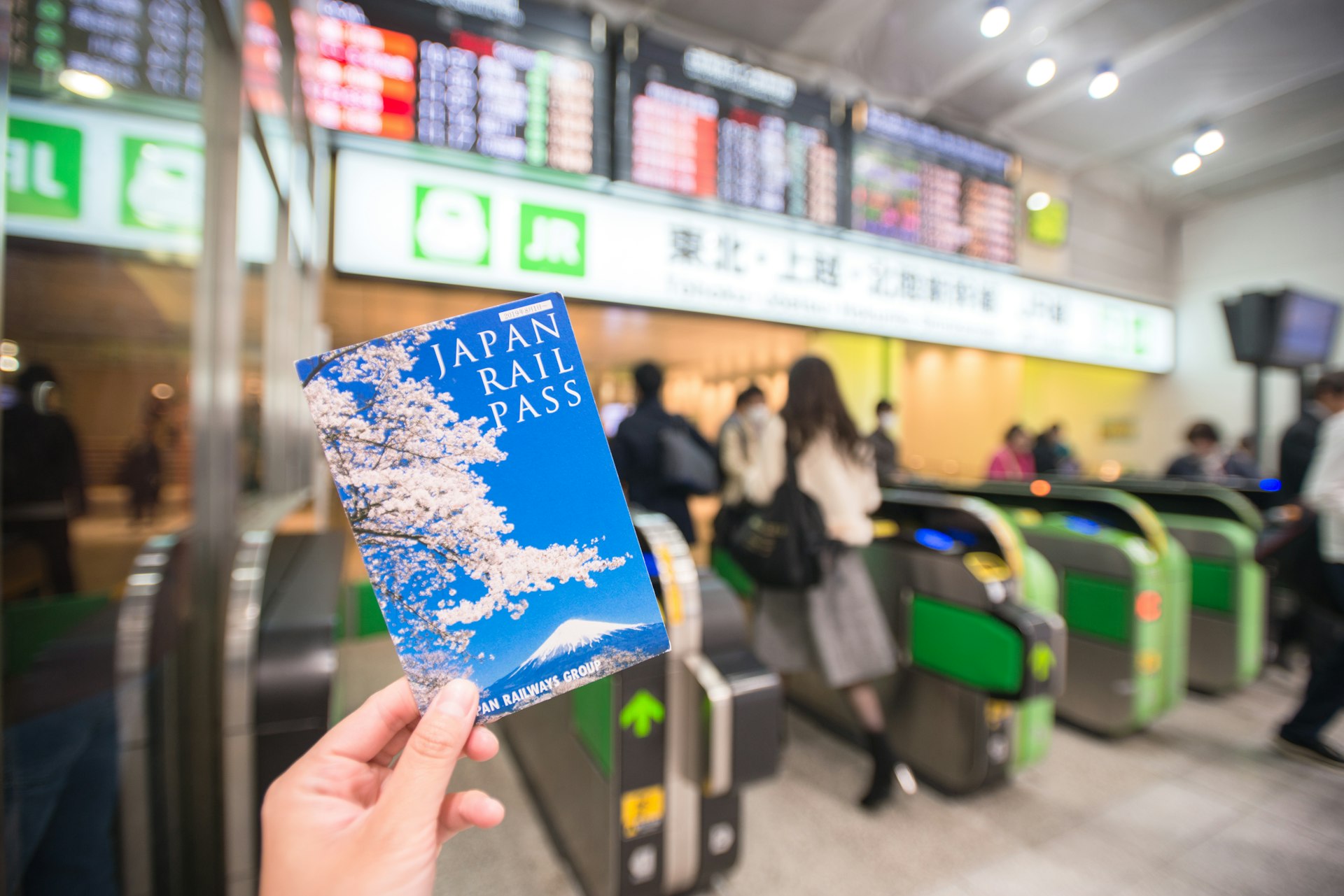 A hand holds up a passport-sized document labeled as Japan Rail Pass in front of barriers at a Japanese train station