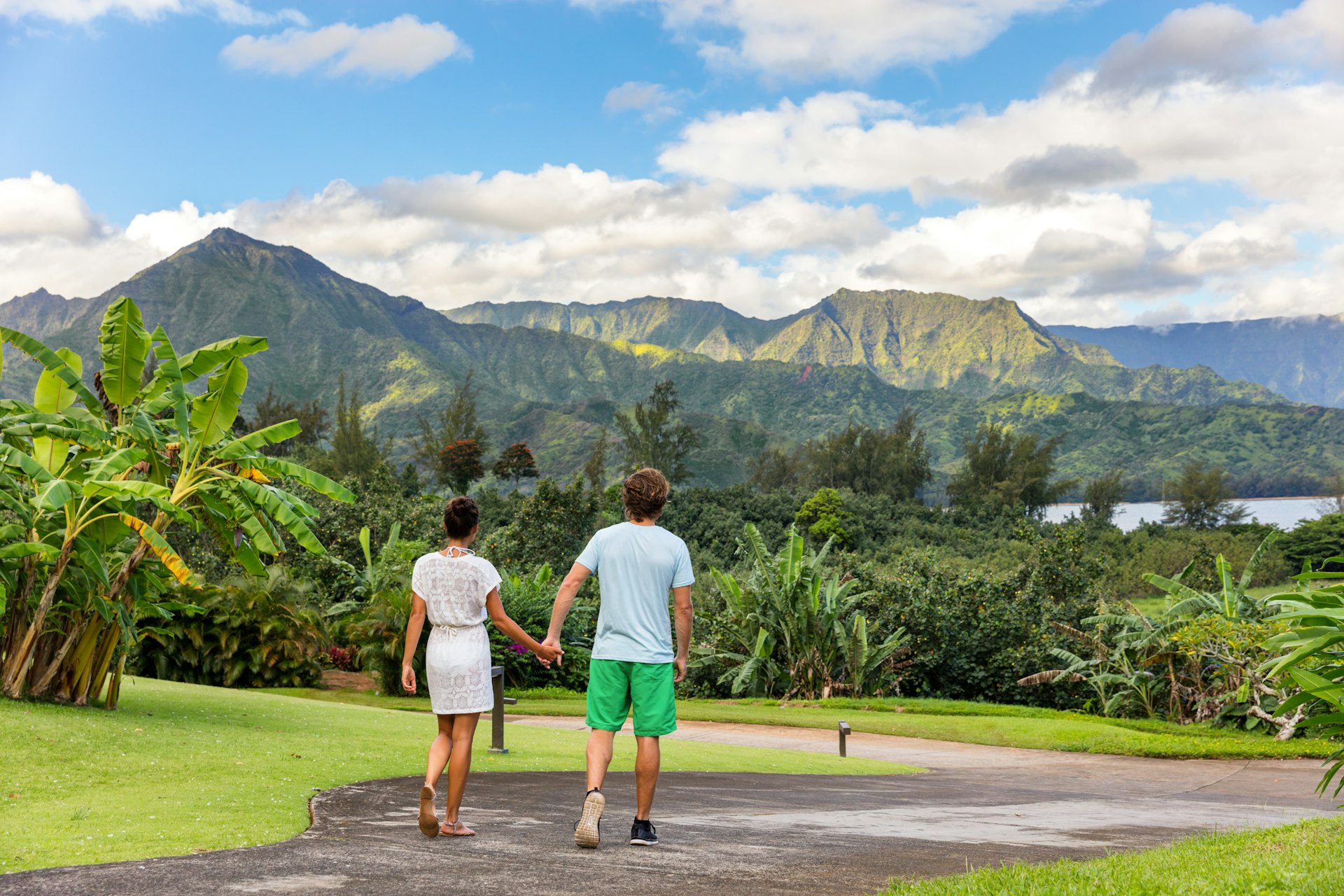A couple walking along a paved pathway surrounded by lush trees, with mountains in the background