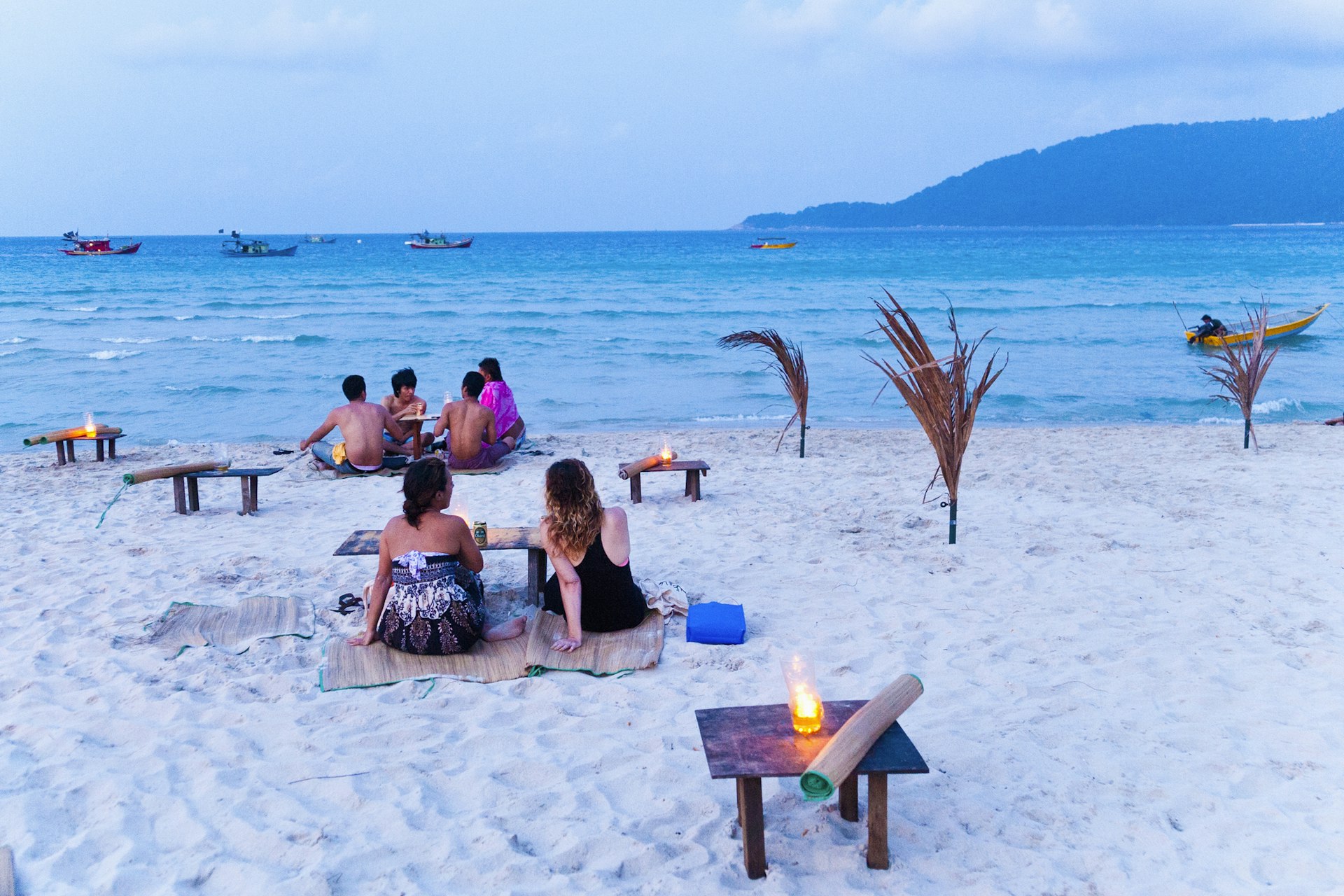 Tourists sit on a beach at dusk. The sand is white and the sea is turquoise. Lanterns have been lit as the light starts to fade. 