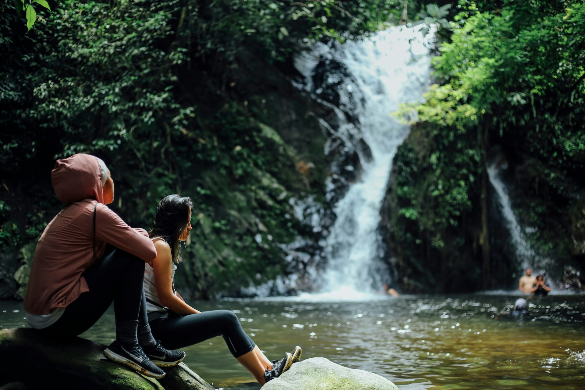 Two women sit by the side of a pool near a waterfall in a jungle