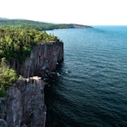 Palisade Head, a towering rock face topped with pines and other vegetation, overlooking Lake Superior on a clear day. Part of Tettegouche State Park
