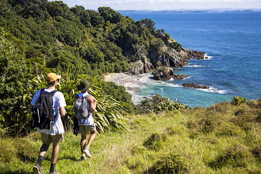 Two hikers wearing backpacks stride down a grassy path towards a sandy cove hidden by tall rocks on each side