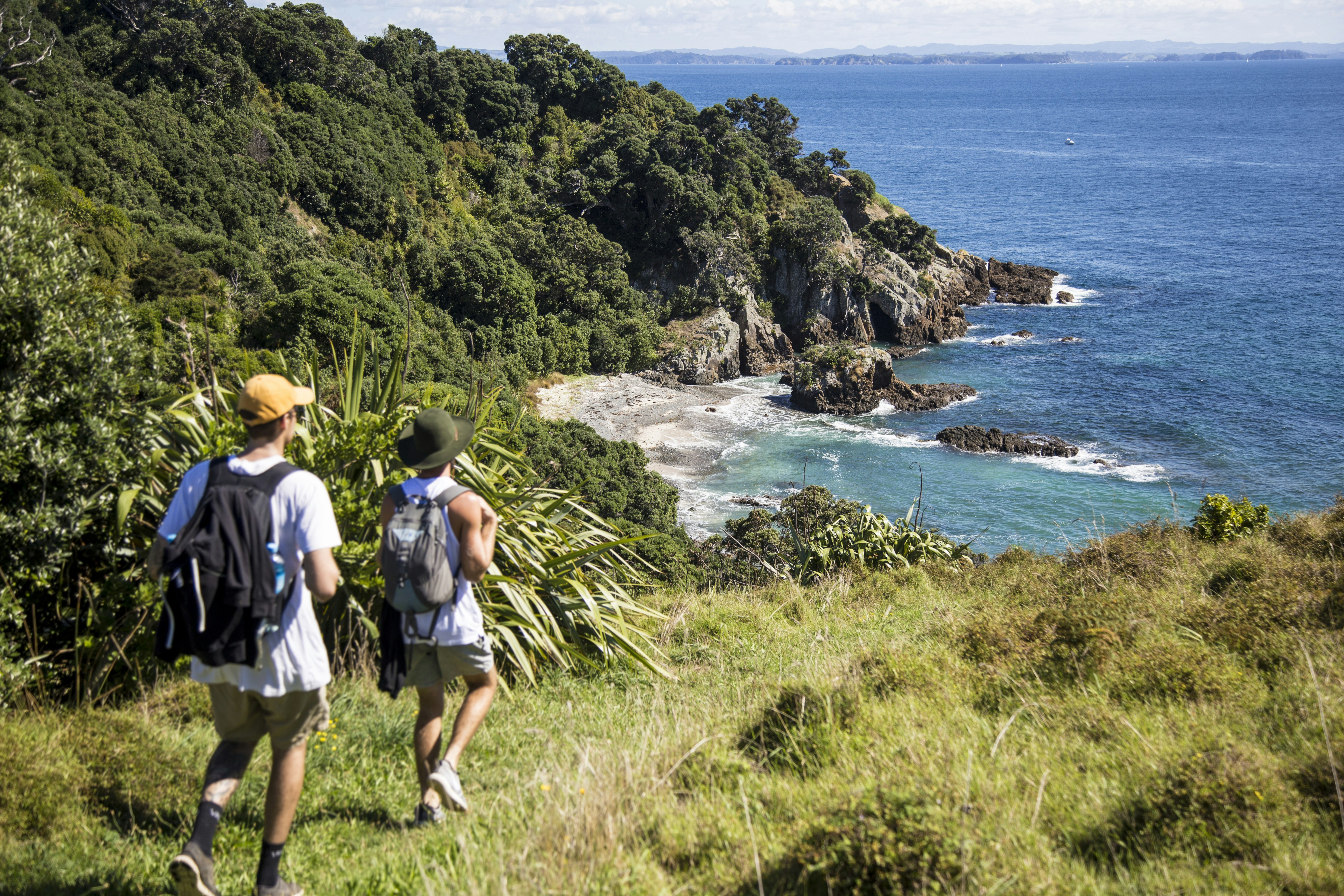 Two hikers wearing backpacks stride down a grassy path towards a sandy cove hidden by tall rocks on each side
