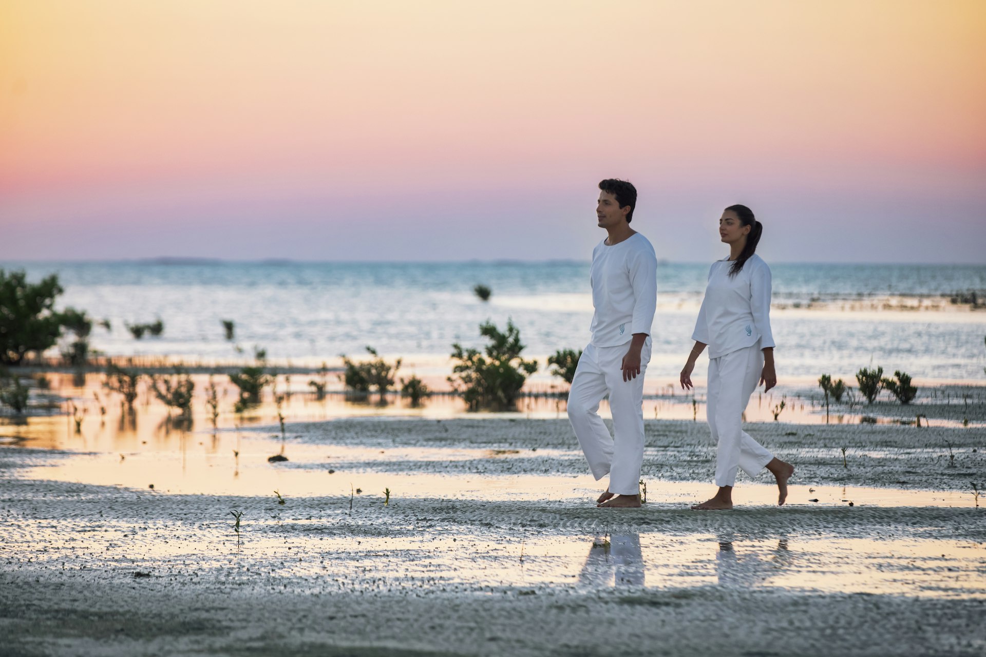 A couple, a man and a woman in white shirt and trousers, walk along a beach in Qatar as the sun sets in orange and purple behind them as part of the Zulal Wellness Resort.