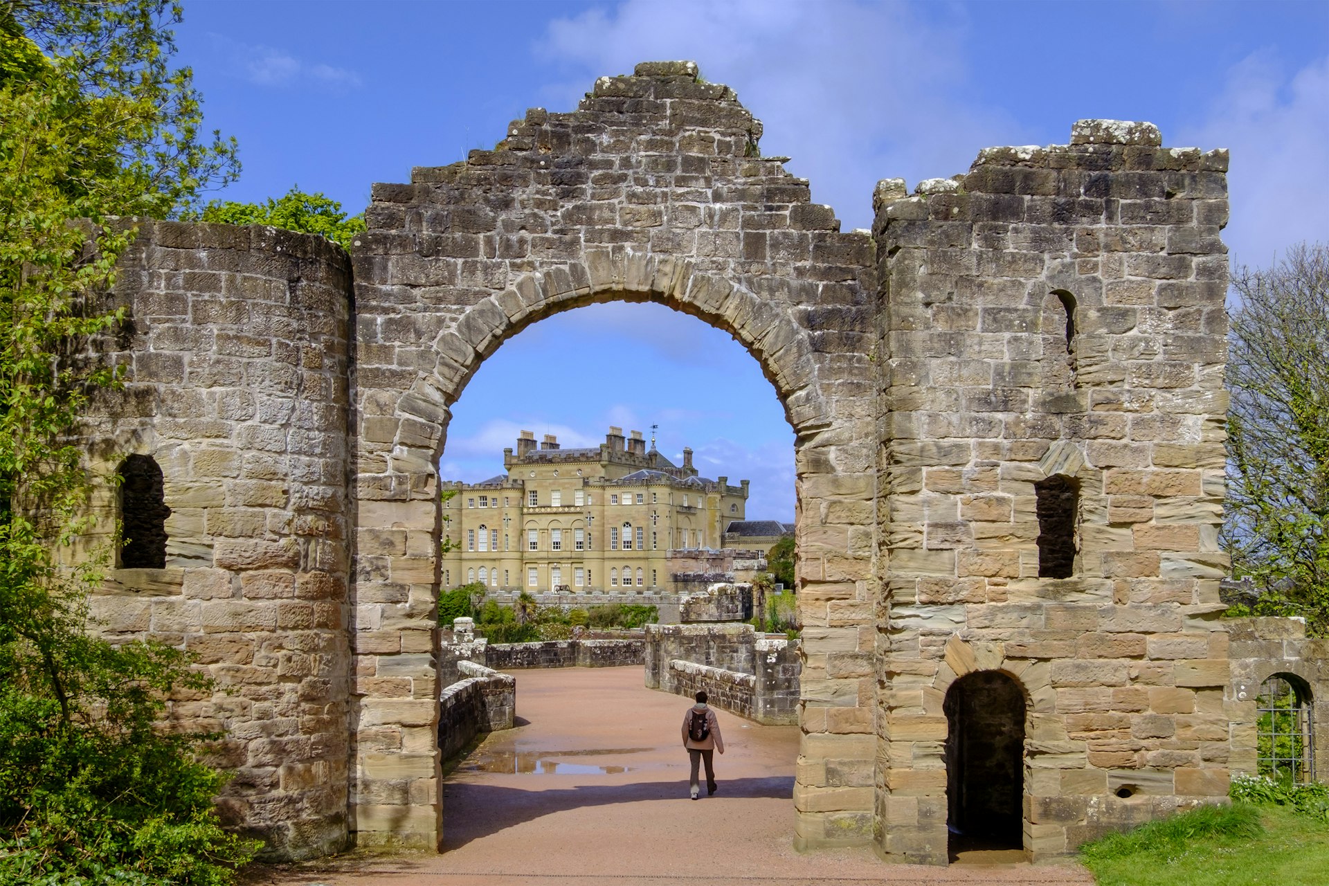 A solo figure walking away from the camera is dwarfed by the giant stone archway over the path. A castle is framed by the arch