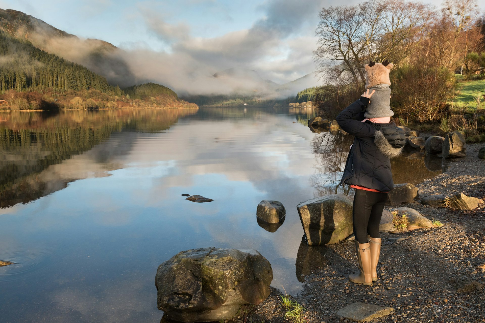 Mother carrying baby on her shoulders glances at misty Loch Lomond in the Trossachs National Park, Scotland, UK