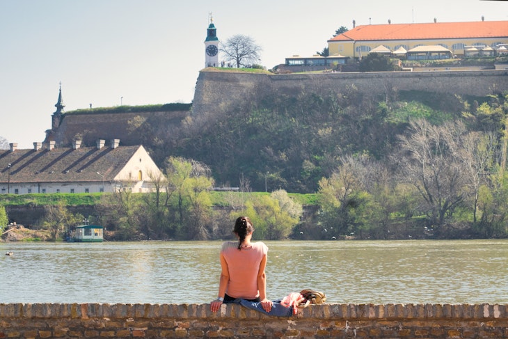 A young woman sitting on the banks of the Danube river in Novi Sad, Serbia, and enjoying the view of the old Petrovaradin fortress; Shutterstock ID 1348024769; your: Claire Naylor; gl: 65050; netsuite: Online Ed; full: Novi Sad weekend update
