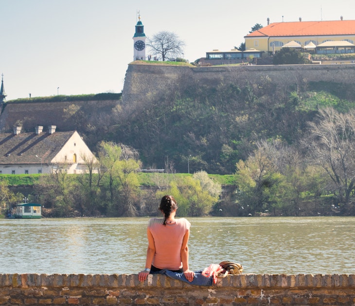 A young woman sitting on the banks of the Danube river in Novi Sad, Serbia, and enjoying the view of the old Petrovaradin fortress; Shutterstock ID 1348024769; your: Claire Naylor; gl: 65050; netsuite: Online Ed; full: Novi Sad weekend update