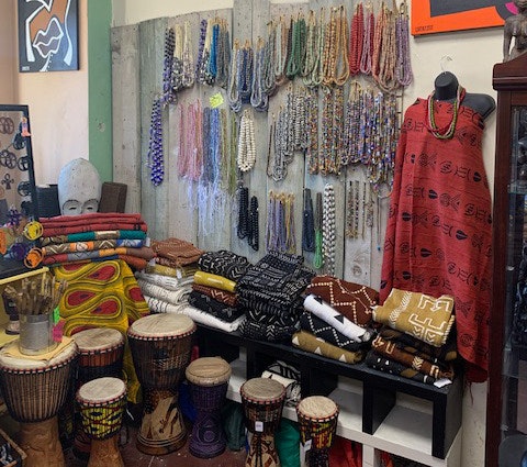 Interior shot of the Sika Jewelry store. There is a large display of necklaces hanging from the wall. Folded fabric and clothing placed on a wooden shelf. A headless mannequin is wearing draped in fabric and there are small drums placed on the floor