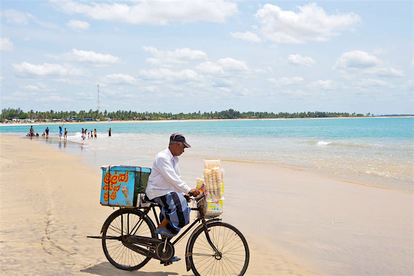 An ice cream seller riding his bike across the sandy beach at Arugam Bay on a bright, partly cloudy day